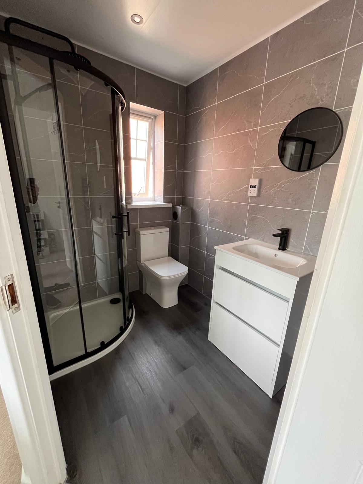Completed shower room with quadrant enclosure, toilet, white vanity unit, round black mirror and grey wood-effect flooring